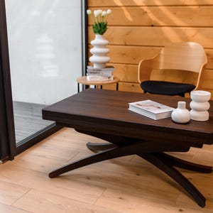 May include: A dark brown wooden coffee table with a unique X-shaped base. The table has a smooth, polished surface and is decorated with two white ceramic vases and a book. The table is set against a backdrop of a light wood wall.