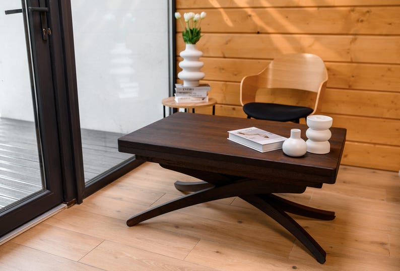 May include: A dark brown wooden coffee table with a unique X-shaped base. The table has a smooth, polished surface and is decorated with two white ceramic vases and a book. The table is set against a backdrop of a light wood wall.