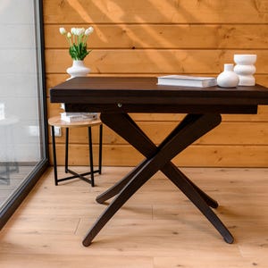 May include: A dark brown wooden table with a black X-shaped base. The table is set against a wood-paneled wall and has a white vase with white flowers on top.