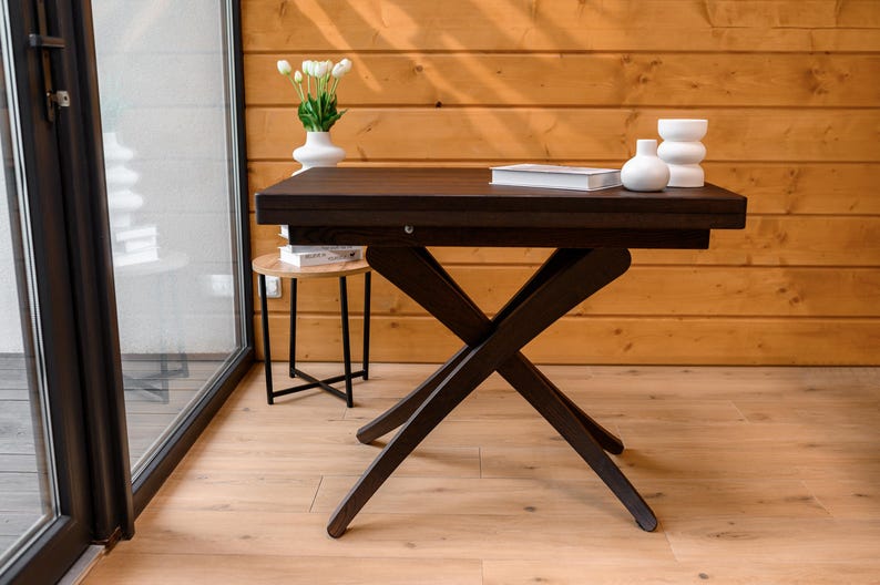 May include: A dark brown wooden table with a black X-shaped base. The table is set against a wood-paneled wall and has a white vase with white flowers on top.