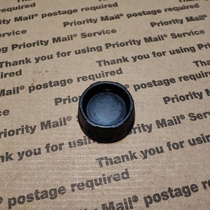 Puede incluir: Un tapón de botella de plástico negro con una parte superior circular y un borde texturizado.