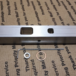 May include: A silver metal tool with a rectangular body, two dials, and two square cutouts. An Allen wrench, a washer, and a small copper ring are also present. The tool is likely used for precision work.