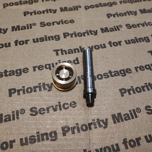May include: A small, circular, gold-toned metal component with a central hole and a separate, slender, dark grey cylindrical metal pin featuring a threaded base. These two distinct hardware pieces are positioned side-by-side on a flat surface.