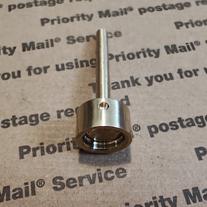 May include: A gold-colored metal bottle stopper with a long, thin stem. The stopper has a ring around the top and a small, round indentation in the center.