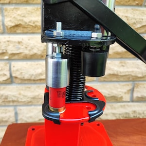 Pode incluir: Uma prensa de recarga vermelha com uma alça preta e uma matriz de recarga prateada. Uma munição de espingarda vermelha está sendo carregada na prensa.