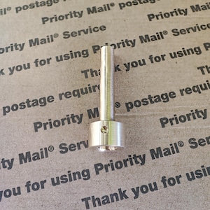 Pode incluir: Um parafuso de metal prateado com cabeça hexagonal e base redonda e lisa. O parafuso está sobre uma superfície marrom com o texto "Priority Mail® Service" impresso nela.