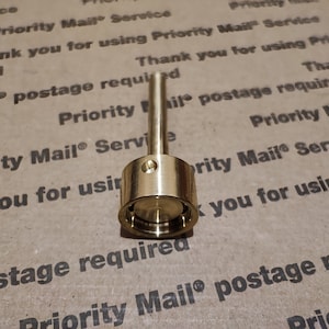 May include: A gold metal bottle stopper with a long, thin stem. The stopper has a small hole in the top and a groove around the bottom.