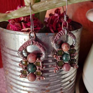 May include: Handmade earrings featuring a unique design. Each earring has a wire-wrapped hoop with a woven brown cord. The earrings are adorned with pink, green, and clear beads. The earrings hang from silver-colored ear wires.