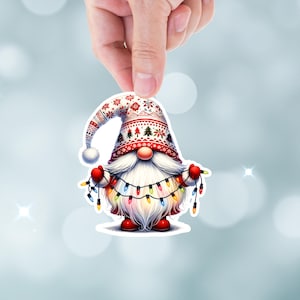May include: A white and red gnome with a white beard and a red hat with a white and red pattern. The gnome is holding a string of colorful Christmas lights.