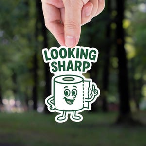 May include: A white sticker with a cartoon toilet paper roll character, smiling and pointing up. The text "LOOKING SHARP" is above the character in green letters. The sticker is held up against a blurred green background.