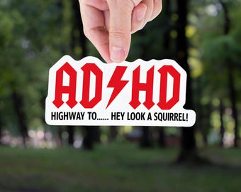 May include: A white sticker with red text that reads "ADHD" with a lightning bolt in the middle, and the text "HIGHWAY TO... HEY LOOK A SQUIRREL!". The sticker is held up by a hand against a blurred green background.