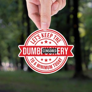 May include: A round, red and white sticker with the text "LET'S KEEP THE DUMB CENSORERY TO A MINIMUM TODAY." The sticker is held up by a hand against a blurred green background of trees.