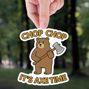 May include: A sticker featuring a brown bear holding an axe, with the text "CHOP CHOP" above and "IT'S AXE TIME" below. The bear has a serious expression. The sticker is held by a hand against a blurred forest background.