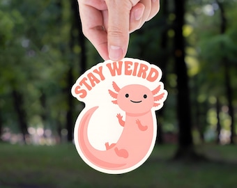 May include: A sticker featuring a cartoon axolotl in shades of pink, with the words "STAY WEIRD" in a coral color. The axolotl has a smiling face and is set against a blurred green background.
