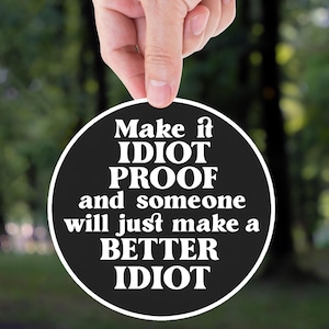 May include: A black and white circular sign with the text "Make it IDIOT PROOF and someone will just make a BETTER IDIOT" is held up against a blurred green forest background. The sign is held by a hand.