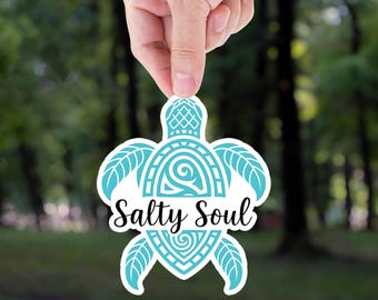 Puede incluir: Una pegatina blanca con forma de tortuga marina, sostenida por una mano. El diseño de la tortuga es turquesa con patrones intrincados y las palabras "Salty Soul" en una fuente de escritura. El fondo es un bosque borroso.