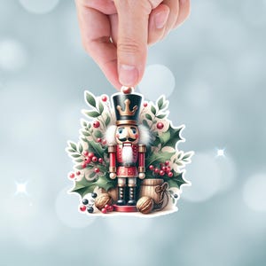 May include: A Christmas nutcracker figurine with a red and gold uniform, a black hat with a gold crown, and a white beard. The nutcracker is surrounded by green holly leaves and red berries.