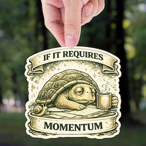 May include: A sticker featuring a cartoon turtle with a coffee mug, resting on a stack of papers. The sticker has the text "IF IT REQUIRES MOMENTUM" in a banner design. The sticker is held up by a hand against a blurred green background.