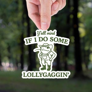 Sticker-style illustration of a frog standing upright in cowboy boots, overalls, and a hat, with a confident stance and a long tongue sticking out. Above and below the frog, green text reads “Y’all mind if I do some LOLLYGAGGIN’” in a mix of playful and bold fonts, giving the design a humorous, Southern-inspired charm.
