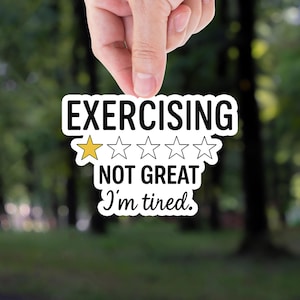 May include: A white sticker with black text that reads "EXERCISING NOT GREAT I'm tired." The sticker has a rating of one gold star out of five. The sticker is held by a hand against a blurred green forest background.