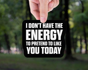 May include: A black rectangular sign with white text that reads "I DON'T HAVE THE ENERGY TO PRETEND TO LIKE YOU TODAY." The sign is held up against a blurred green background of trees.