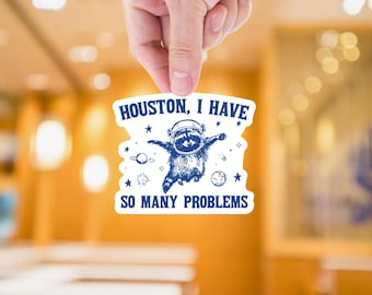 May include: White sticker with blue text and a cartoon raccoon wearing a space helmet. The text reads "Houston, I have so many problems".