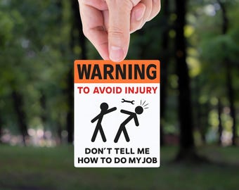 May include: A rectangular sign with an orange top and white bottom, held by a hand. The sign reads "WARNING TO AVOID INJURY" and depicts stick figures. The text "DON'T TELL ME HOW TO DO MY JOB" is also present.