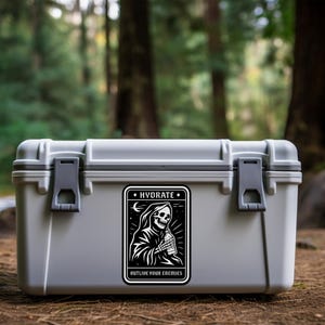 May include: A gray, rectangular cooler with a black and white sticker featuring a Grim Reaper holding a water bottle. The sticker reads "HYDRATE" and "OUTLIVE YOUR ENEMIES". The cooler is outdoors.