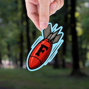 May include: A red bomb sticker with the letter "F" in black, held by a hand against a blurred forest background. The sticker has a light blue outline and stylized motion lines.