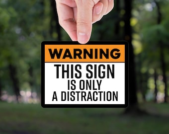 May include: A black rectangular sign with an orange header that reads "WARNING". The sign continues with the text "THIS SIGN IS ONLY A DISTRACTION" in bold white letters. The sign is held up by a hand against a blurred green background.
