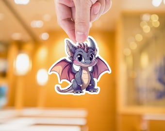 May include: A sticker featuring a cartoon dragon with a friendly expression. The dragon is primarily gray with pink wings and accents. It has large, expressive eyes and is held up by a hand against a blurred background.