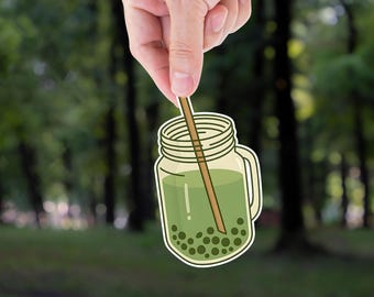 May include: A sticker of a mason jar filled with green liquid and dark green boba pearls. A brown straw is inside the jar. The jar has a handle and a screw-on lid. The background is a blurred image of a forest.