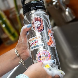 May include: A stainless steel water bottle with a black handle and a variety of colorful stickers, including a baseball glove, a donut, a dog, and a quote that says "Comfort is the enemy of achievement".