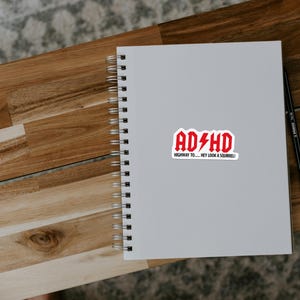 May include: A white spiral-bound notebook with a red and white sticker that reads "ADHD" with a lightning bolt graphic. The sticker also includes the text "HIGHWAY TO... HEY LOOK A SQUIRREL!" A black pen rests on the right side.