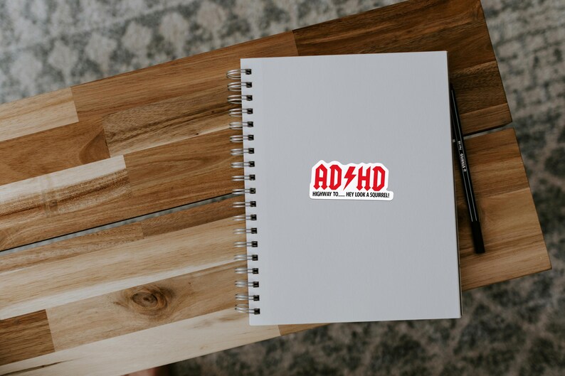 May include: A white spiral-bound notebook with a red and white sticker that reads "ADHD" with a lightning bolt graphic. The sticker also includes the text "HIGHWAY TO... HEY LOOK A SQUIRREL!" A black pen rests on the right side.