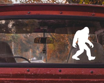 May include: A white silhouette of Bigfoot walking, applied as a decal on the rear window of a red pickup truck.