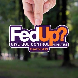 May include: A sticker with the text "Fed Up?" in white and orange, with the words "GIVE GOD CONTROL HE DELIVERS" and "Psalm 34:19" below. The sticker is held by a hand against a blurred green background.