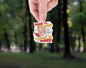 May include: A sticker with a vintage comic book style design. The sticker features a distressed blonde woman reading a book, with text that reads "Born to Read, Forced to Work". The background is a blurred green forest.
