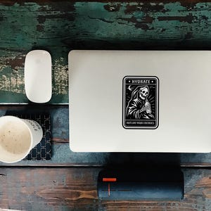 May include: A white laptop with a black and white sticker that says "HYDRATE" with a grim reaper design. The laptop is on a distressed wooden surface with a white mouse, a cup of coffee, and a black speaker.