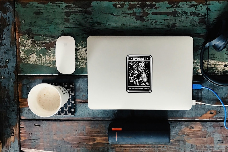 May include: A white laptop with a black and white sticker that says "HYDRATE" with a grim reaper design. The laptop is on a distressed wooden surface with a white mouse, a cup of coffee, and a black speaker.