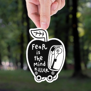 May include: A black and white sticker in the shape of an apple, held by a hand. The sticker features the text "Fear is the Mind Killer" and a cartoon figure inside. The background is a blurred forest.