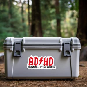 May include: A gray, rectangular storage container with black latches and a red and white sticker that reads "ADHD Highway to... Hey look a squirrel!" The container is outdoors, set against a blurred forest background.