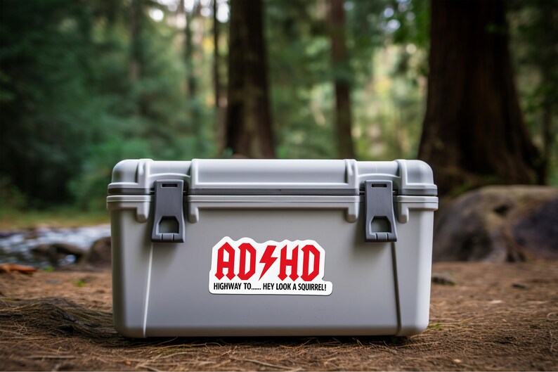 May include: A gray, rectangular storage container with black latches and a red and white sticker that reads "ADHD Highway to... Hey look a squirrel!" The container is outdoors, set against a blurred forest background.
