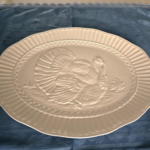 May include: An oval, off-white serving platter with a raised relief of a turkey. The platter has a decorative border with a ribbed edge and a cross-hatch pattern. The turkey is depicted with its feathers fanned out.