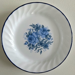 Pode incluir: Prato de cerâmica branco com um desenho floral azul no centro. O prato tem uma borda azul escura e um padrão em espiral na borda. O desenho floral apresenta rosas azuis e outras flores.