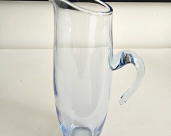 May include: A tall, light blue glass pitcher with a curved handle. The pitcher has a slender, elegant shape and a pouring spout. The glass is transparent, allowing light to pass through. Ideal for serving beverages.