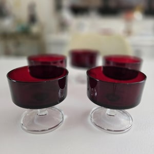 May include: Five stemmed glasses with a deep red interior and a clear, rounded base. The glasses have a dark, translucent exterior, and are arranged on a white surface. The focus is on the front two glasses.