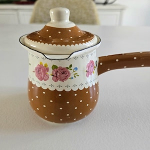 Op de afbeelding: Een bruine en witte emaille steelpan met een deksel. De steelpan heeft een bruin stippenpatroon op de bodem en het deksel. Het bovenste deel van de steelpan is wit met roze rozenbloemmotieven. De handgreep is bruin met witte stippen.