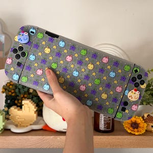 Peut inclure: Une coque grise pour console Nintendo Switch avec un motif répété de fruits et d'étoiles colorés. L'étui a des couvre-boutons et des breloques décoratives assortis. La console est tenue dans la main d'une personne.