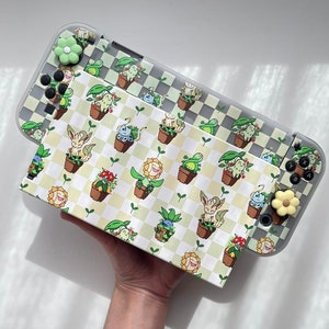 May include: A white Nintendo Switch Lite console with a clear case featuring a checkered pattern and a green and white floral design. The case has small cartoon characters on it. The console is being held by a hand.